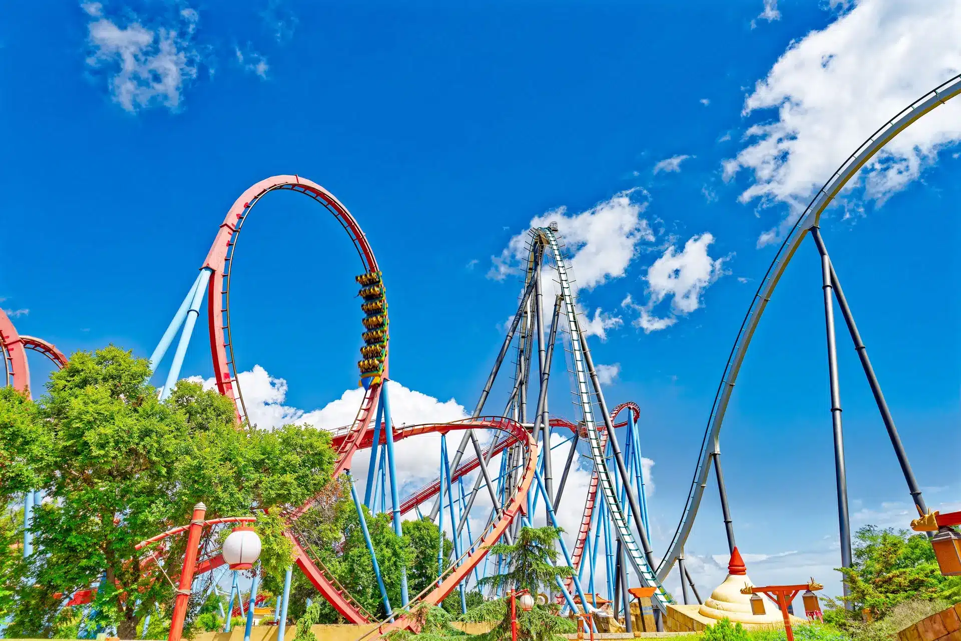 Red and blue roller coaster tracks with a vertical loop and tall supports under a bright blue sky with white clouds