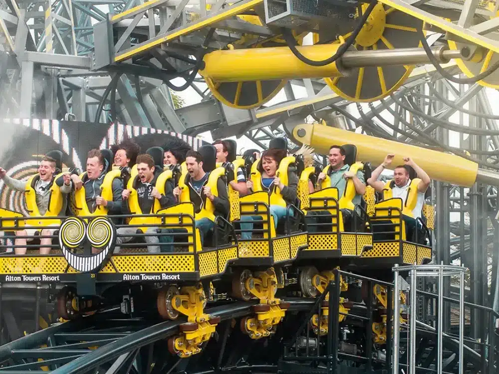 Riders with raised arms on a yellow roller coaster train marked Alton Towers Resort, with a smiling face design on the front