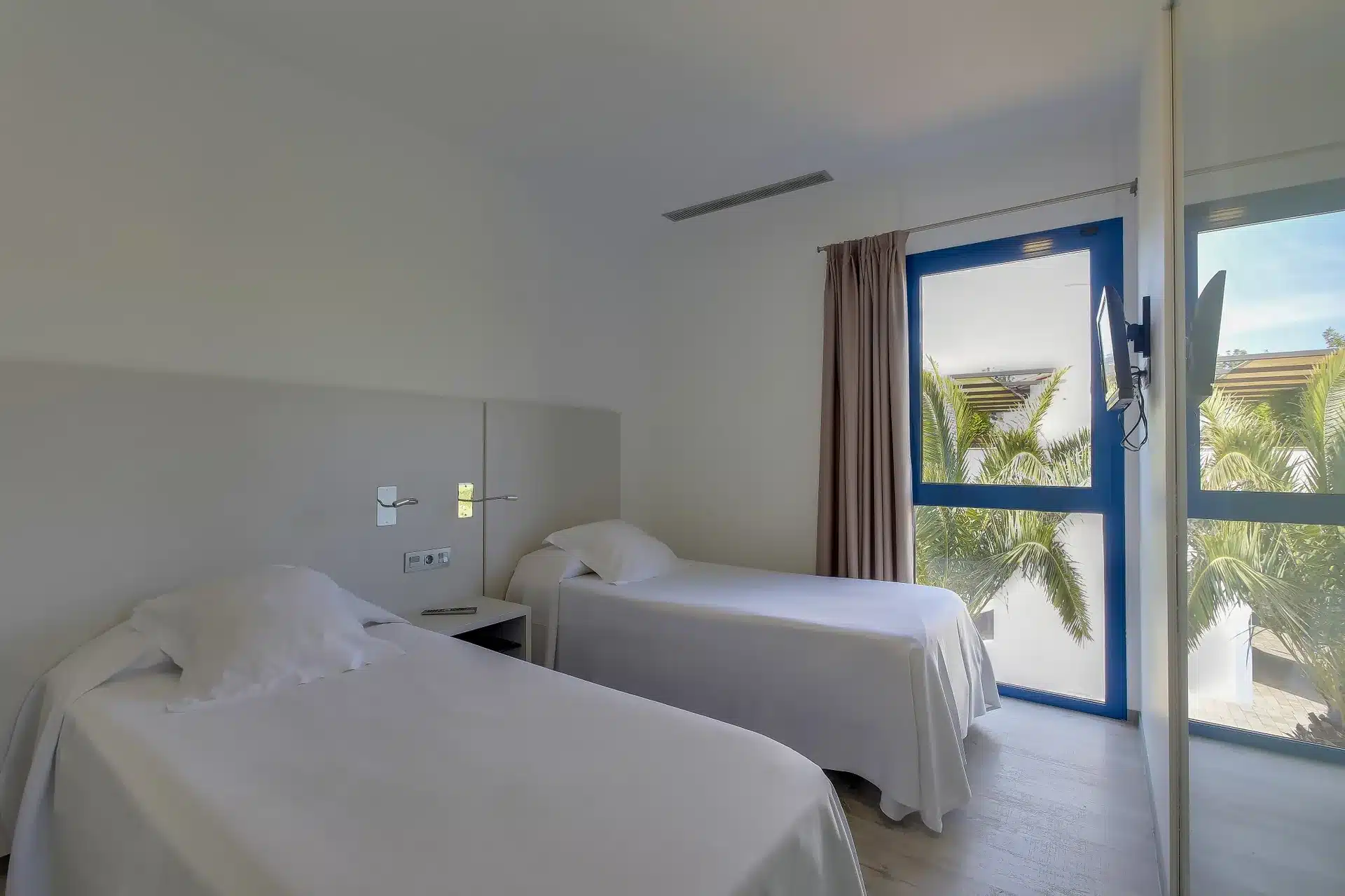 Bedroom with two single beds in white bedding, a wall-mounted TV and a blue-framed glass door looking onto palm trees