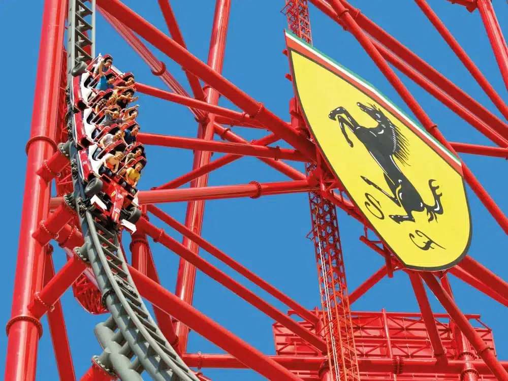 Roller coaster train with riders climbing a steep track beside a large yellow Ferrari shield with a black prancing horse