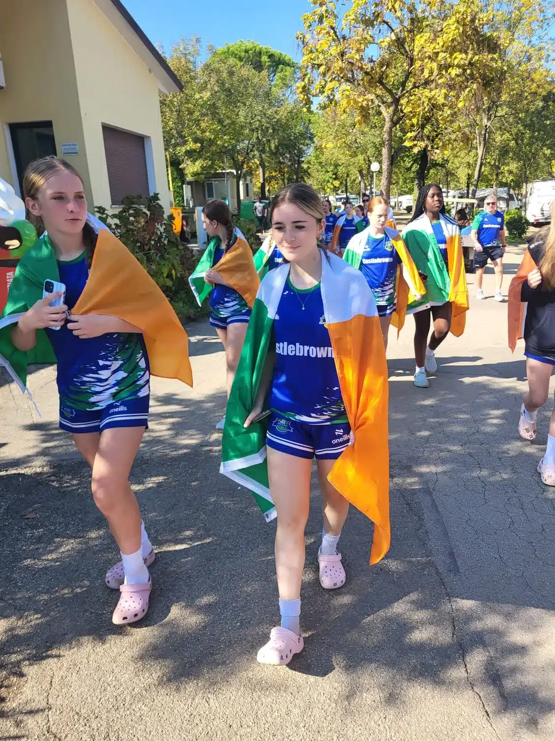 Girl in a blue Castlebrowne jersey with an Irish flag over her shoulders walking on a path, with other girls in the same kit behind her