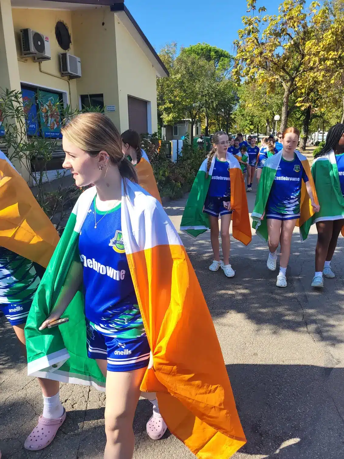 Girls in blue Castlebrowne jerseys walking along a paved path with Irish flags over their shoulders