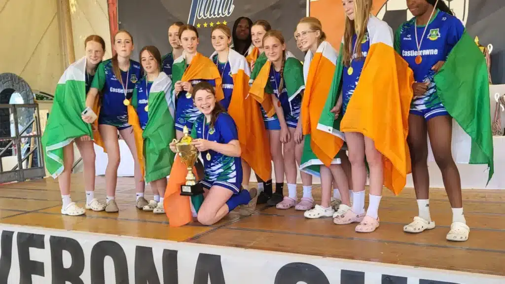 Girls in blue Castlebrowne jerseys draped in Irish flags and wearing medals pose on a stage with a gold trophy, in front of Verona Cup and Bella Italia banners