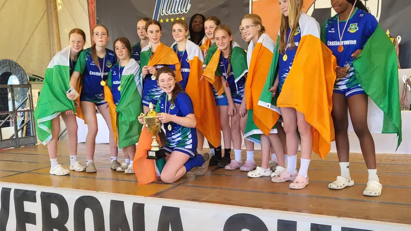 Girls in blue Castlebrowne jerseys draped in Irish flags and wearing medals pose on a stage with a gold trophy, in front of Verona Cup and Bella Italia banners