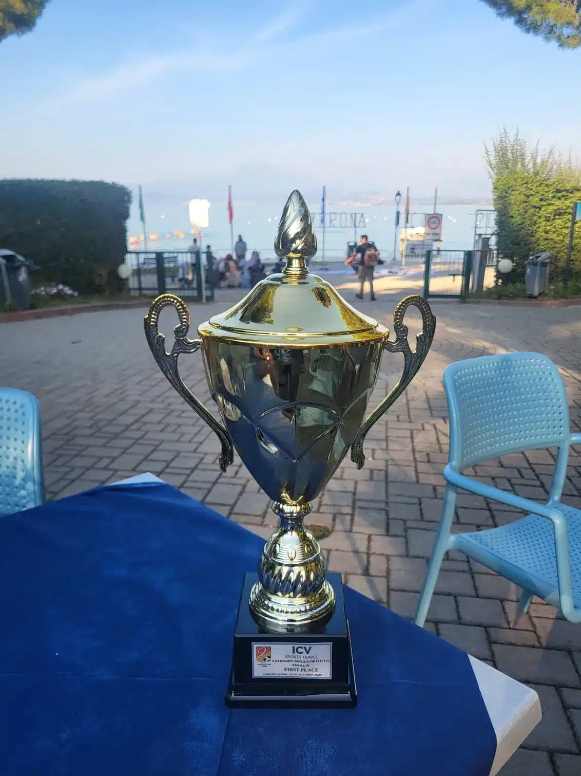 Gold trophy on a blue tablecloth with a plaque reading first place, Category Girls U14, with a lakeside promenade in the background